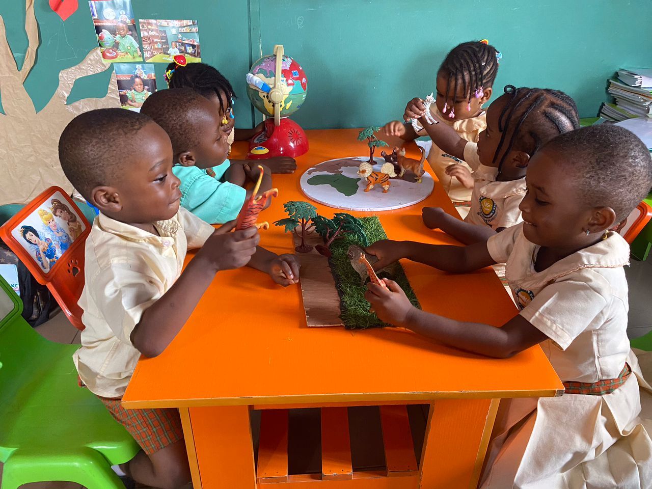 Pupils around an orange classroom table building a handmade model of the African continent with animal figures, beside a globe and a painted map